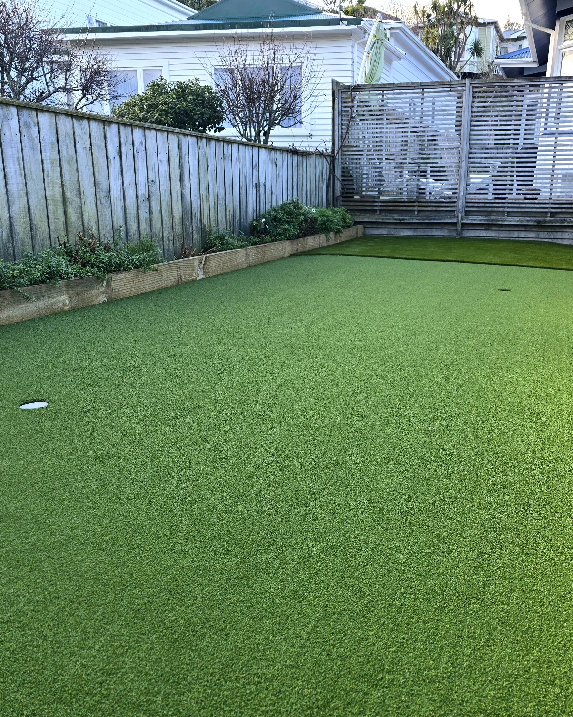 Backyard with a green artificial grass putting green, bordered by a wooden fence and a garden bed with shrubs and small plants, with neighboring houses and trees visible in the background.