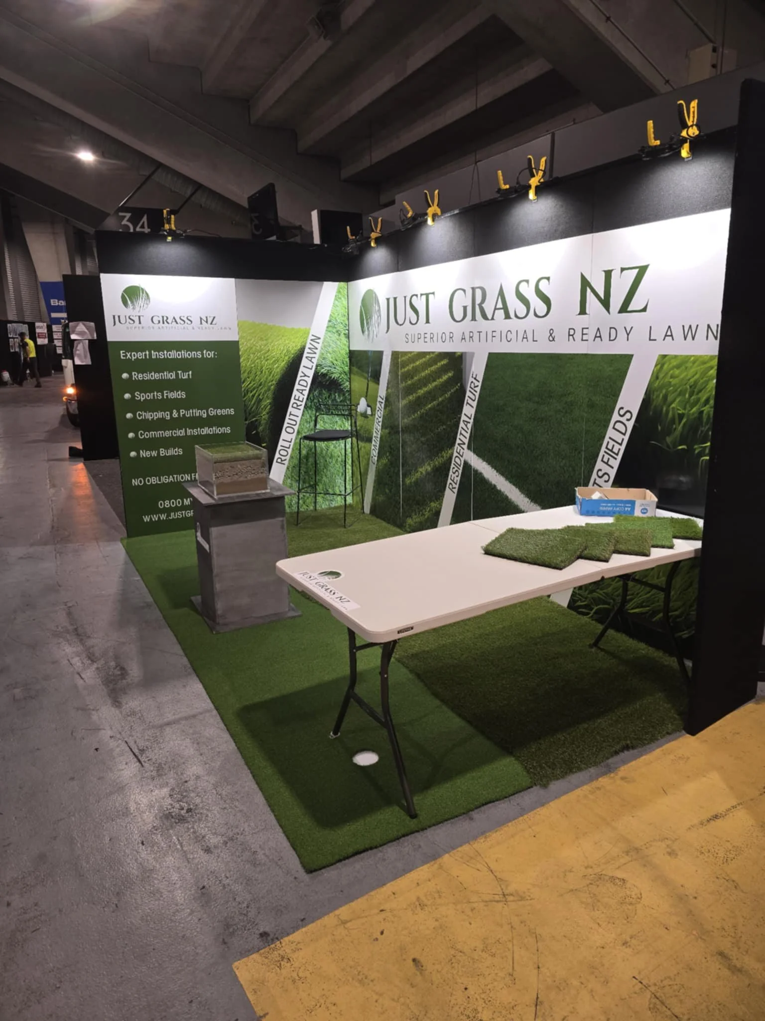 Trade show booth for Just Grass NZ showcasing artificial turf with a table displaying sample turf pieces and a backdrop with green grass images and company branding.