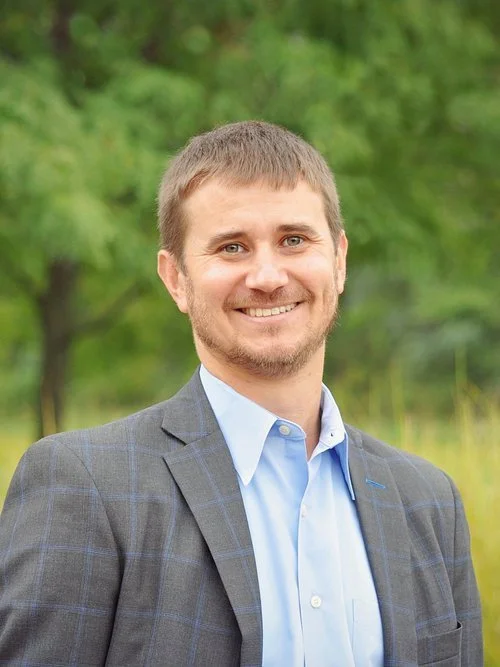A man in a business suit smiling outdoors with trees in the background.