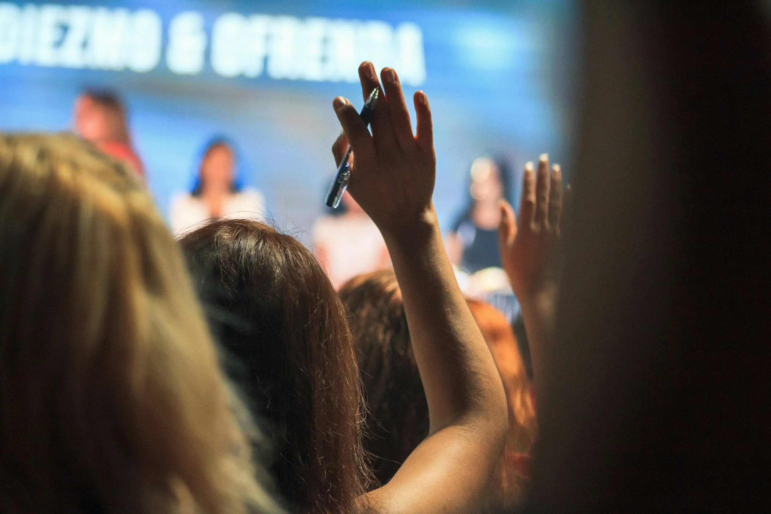 People seated at an event with one person raising their hand and holding a pen or marker, with a blurred background of a screen or presentation.