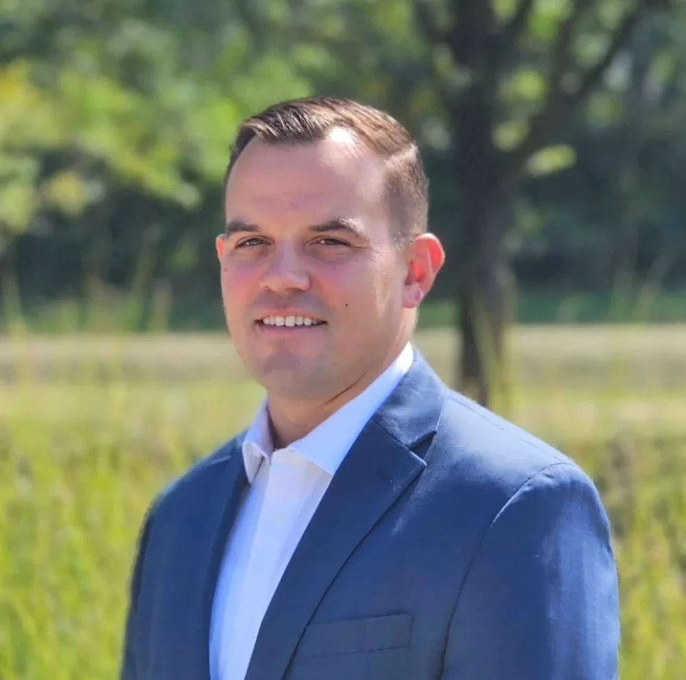 A man in a blue suit and white shirt standing outdoors with trees and grass in the background.
