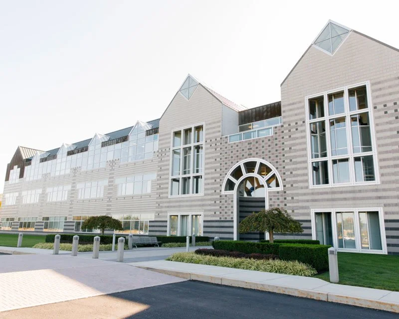Modern multi-story building with large windows and a distinctive arched entrance, surrounded by well-maintained landscaping and a paved walkway.