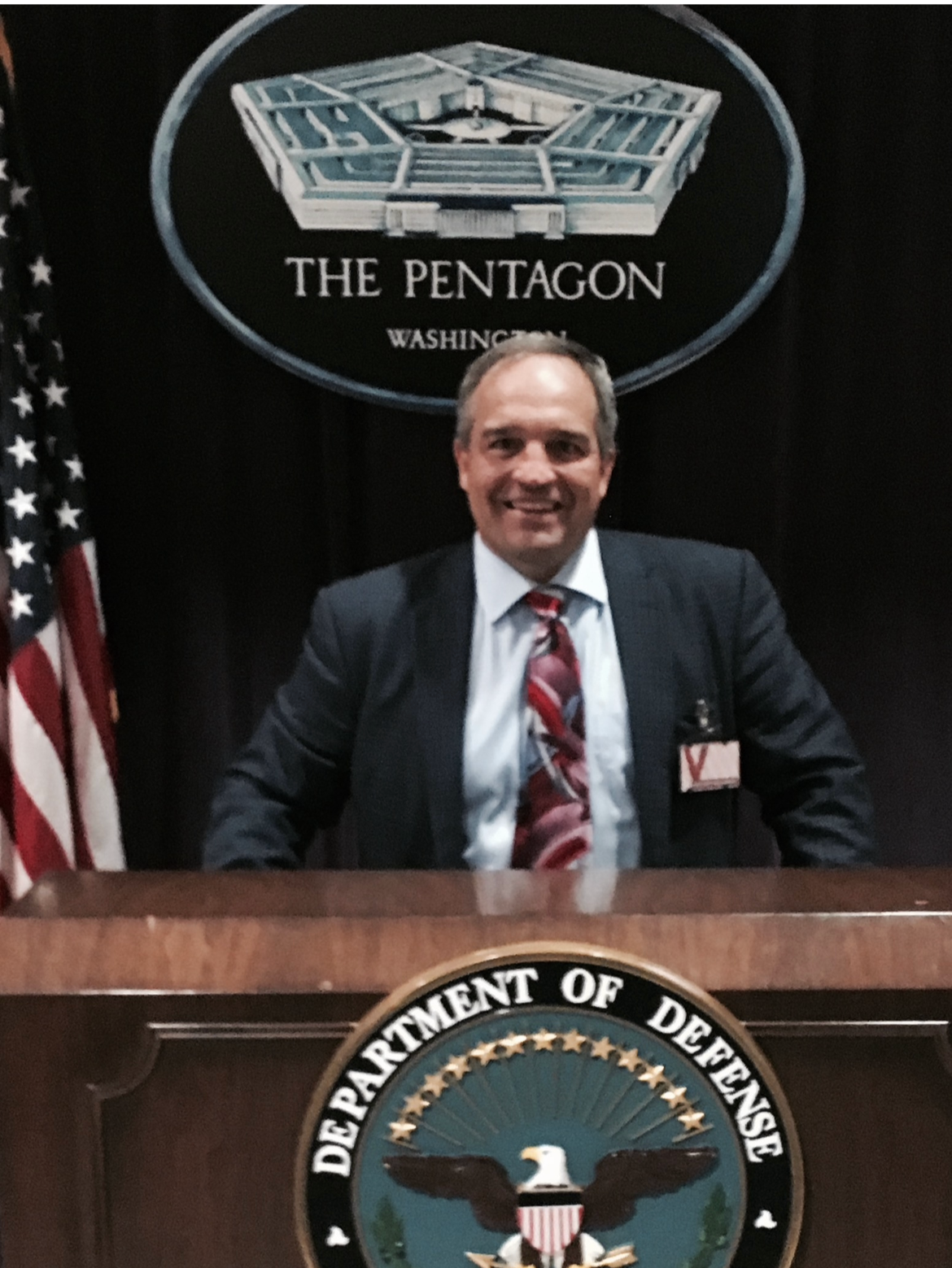 A man sitting at a desk at the Pentagon, Washington, D.C., with an American flag on his left and the Department of Defense seal on the front of the desk. Behind him is a sign with the Pentagon's outline and the words 'The Pentagon, Washington'.