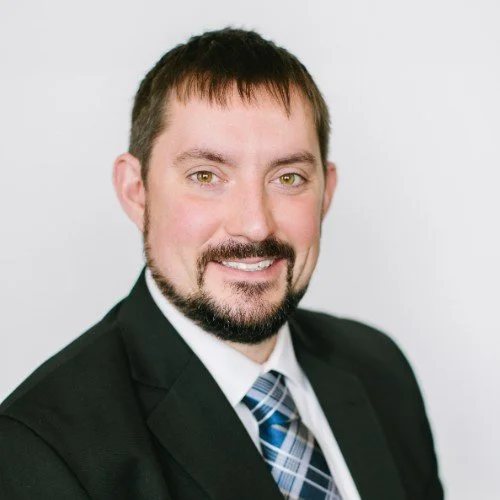 Professional headshot of a man with short brown hair, a beard, wearing a black suit, white shirt, and a blue patterned tie, smiling against a plain white background.