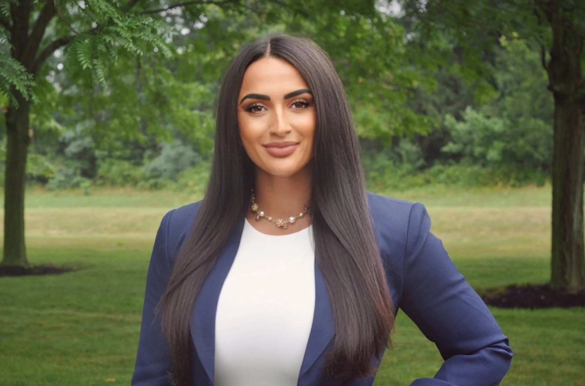A woman with long dark hair, wearing a navy blazer and white top, stands outdoors in a park with green trees in the background.