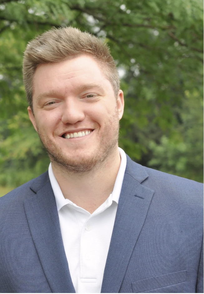 Smiling man in a navy suit outdoors with green trees in the background.
