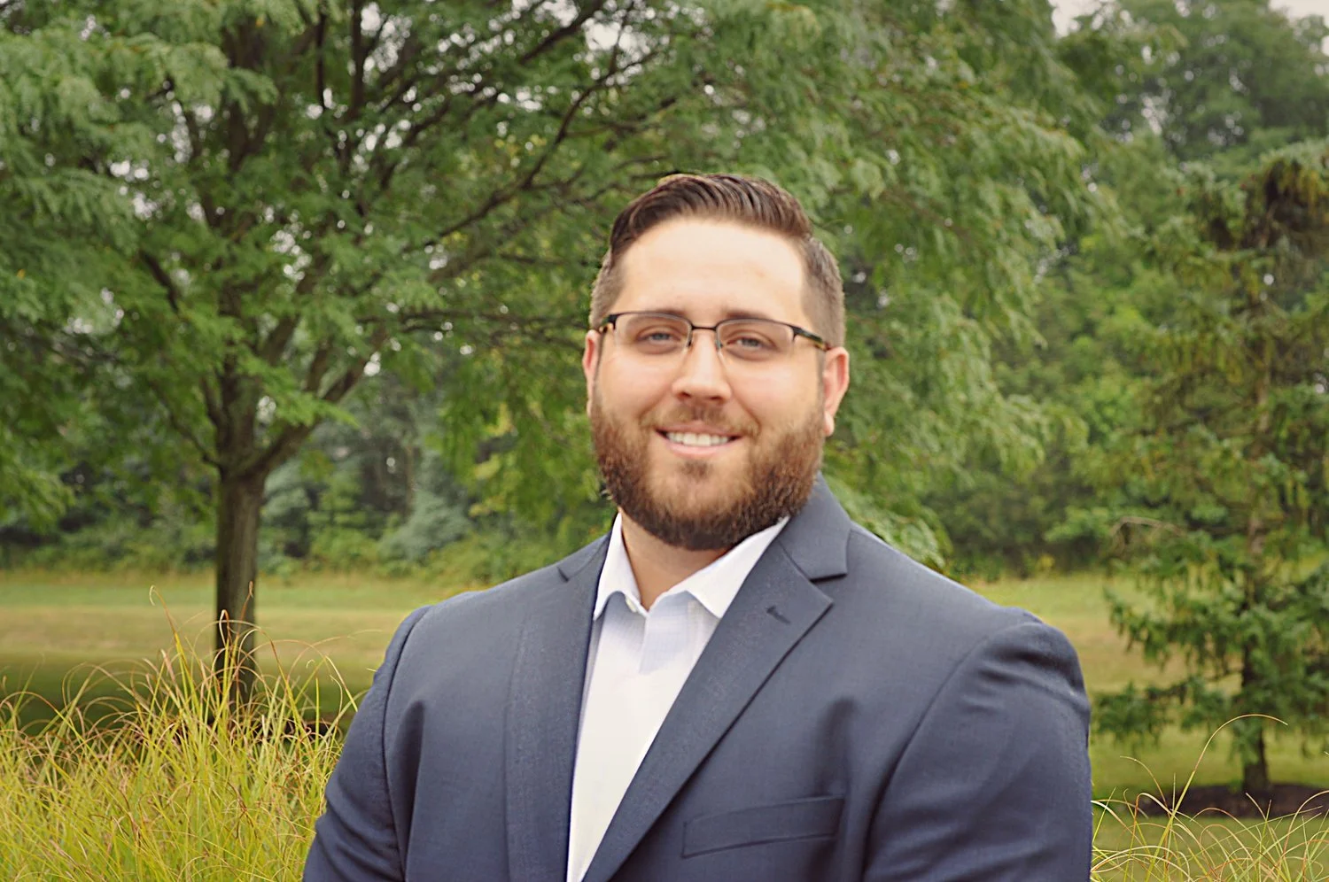 A man with glasses and a beard, wearing a navy suit and white shirt, standing outdoors with trees and grass in the background.