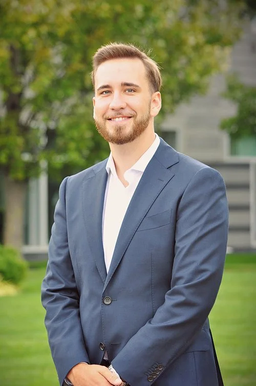 A young man with a beard and mustache wearing a navy blue suit and white dress shirt, standing outdoors on a grassy area with trees and a building in the background.