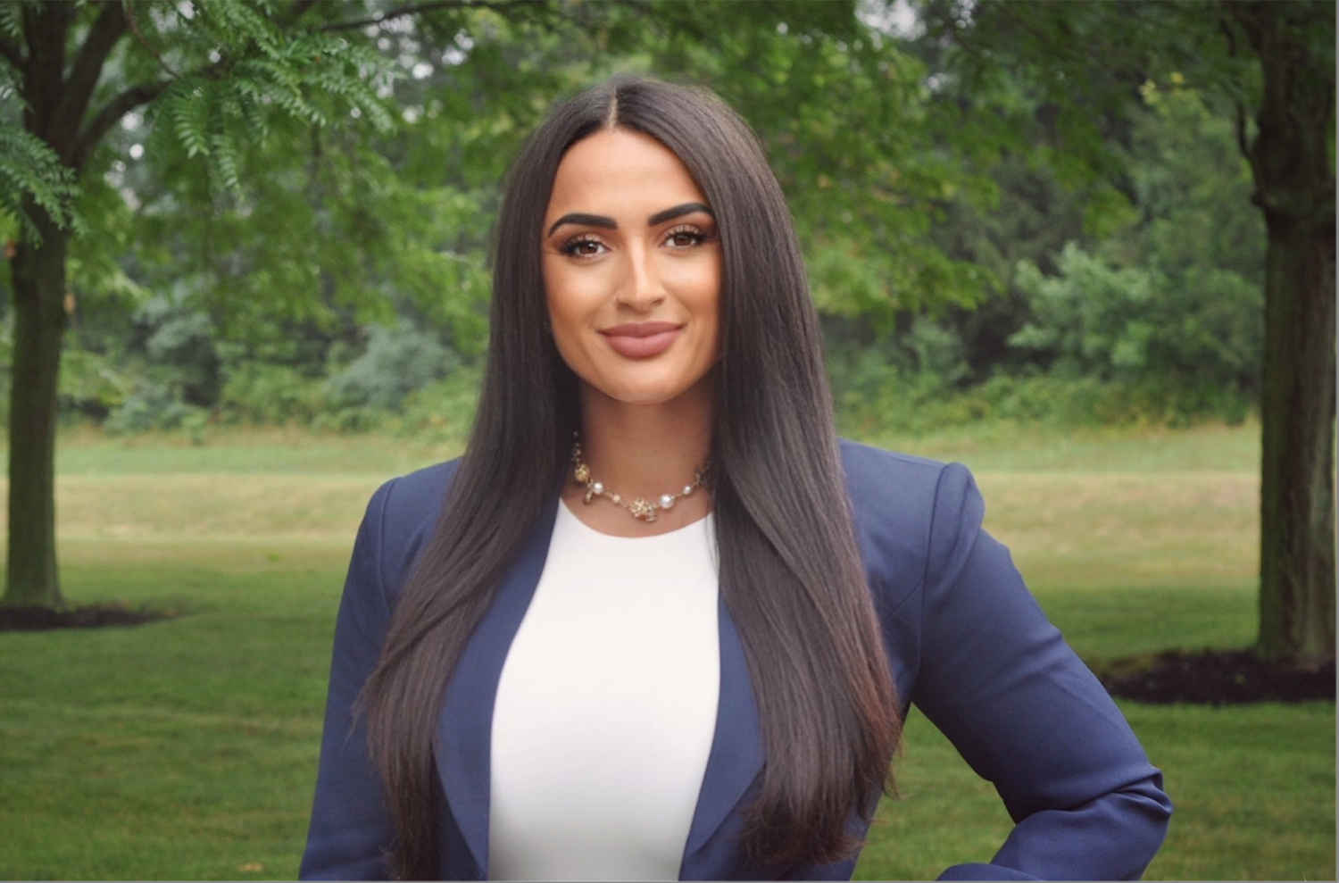 A woman with long dark hair, wearing a navy blazer and white top, standing outdoors in a park with green trees and grass in the background.