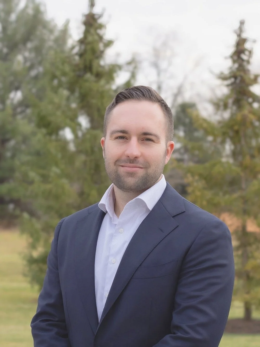 A professional man in a dark suit and white shirt standing outdoors with trees in the background.