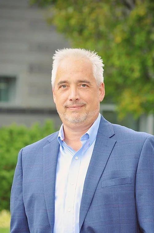 A middle-aged Caucasian man with short white hair and a light beard, wearing a blue checked suit jacket over a light blue dress shirt, standing outdoors with a blurred background of trees and a building.