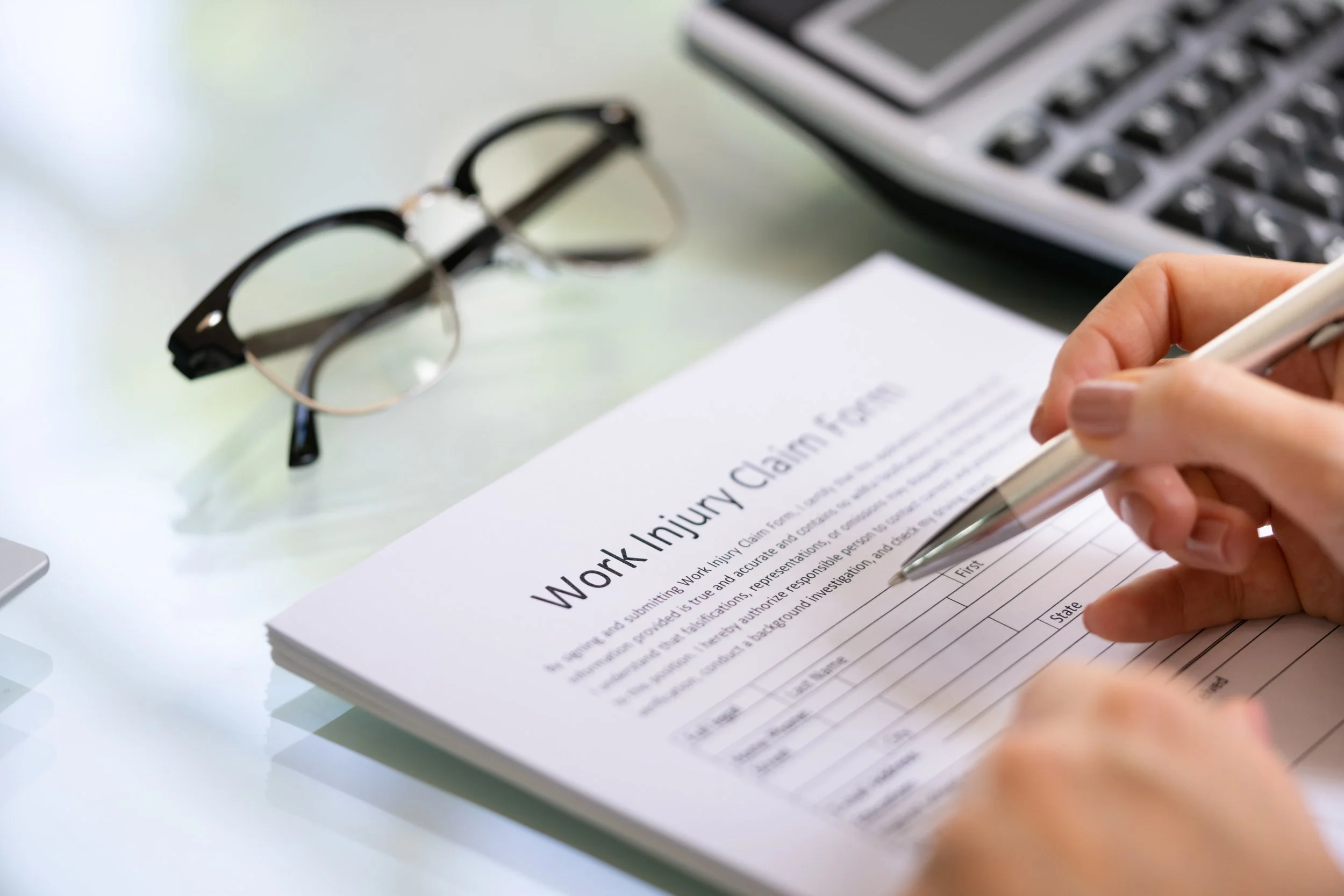 A person filling out a form titled 'Work Injury Claim' with a pen, on a white desk. Eye glasses and a keyboard are nearby.