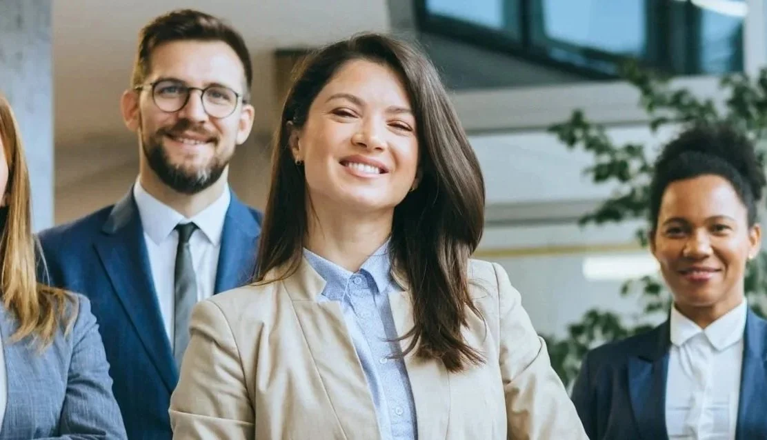 Group of professional diverse women and men in business attire smiling in office setting.