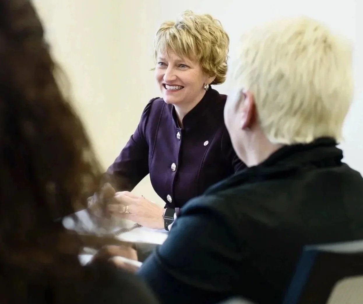 A woman with short curly blonde hair smiling and talking in a meeting with two people, one with blond hair and one with dark hair, all sitting at a table.