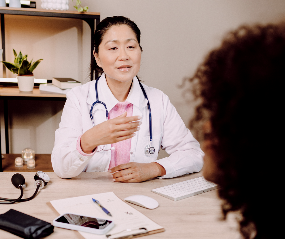 A female doctor in a white coat with a stethoscope around her neck, speaking to a patient in a medical office. The desk has medical equipment, papers, a pen, a smartphone, and a computer keyboard.