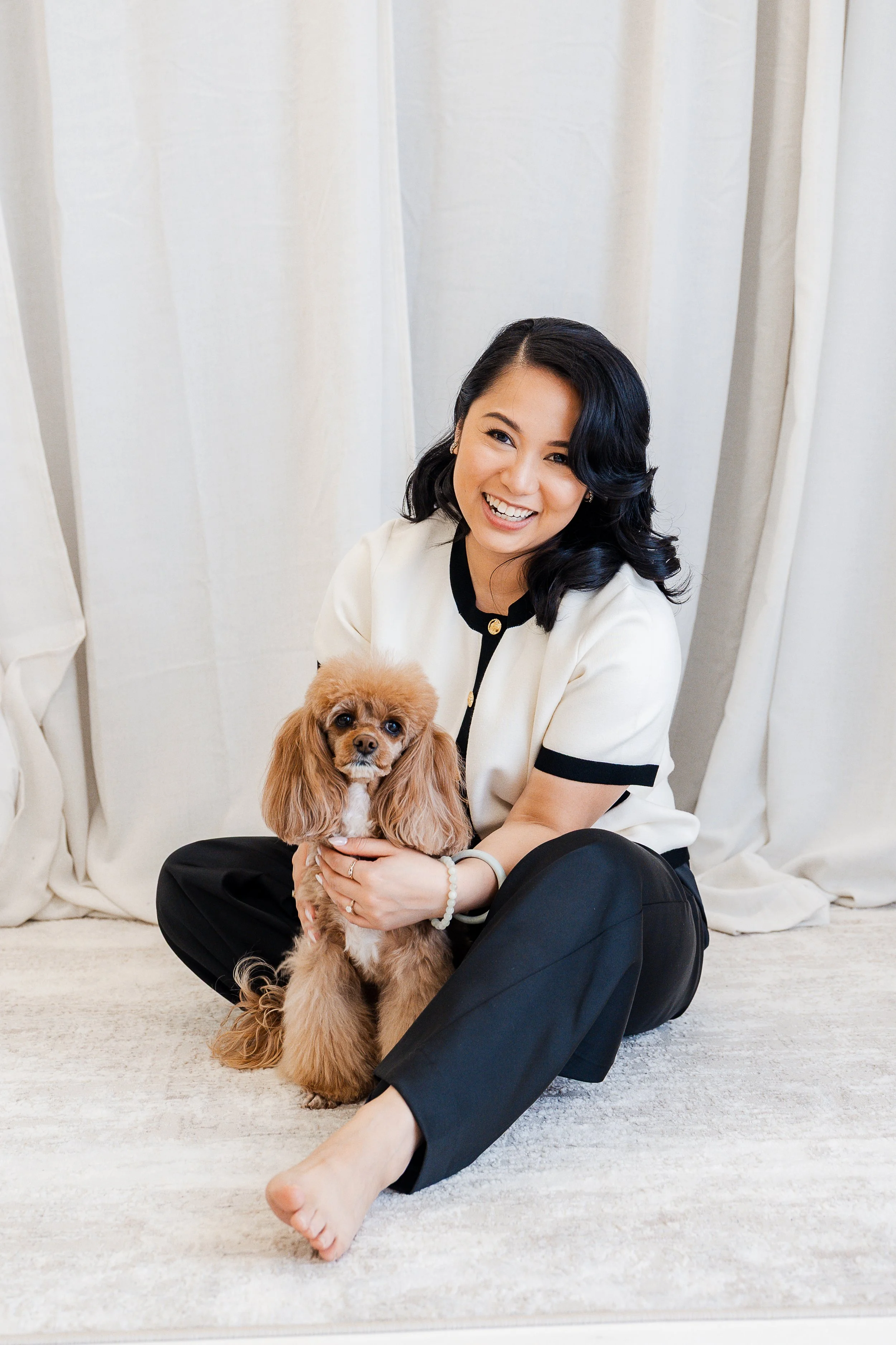 A woman with black hair, wearing a white and black outfit, is sitting on the floor and holding a brown dog with long ears. She is smiling and looking at the camera, with a curtain backdrop behind them.