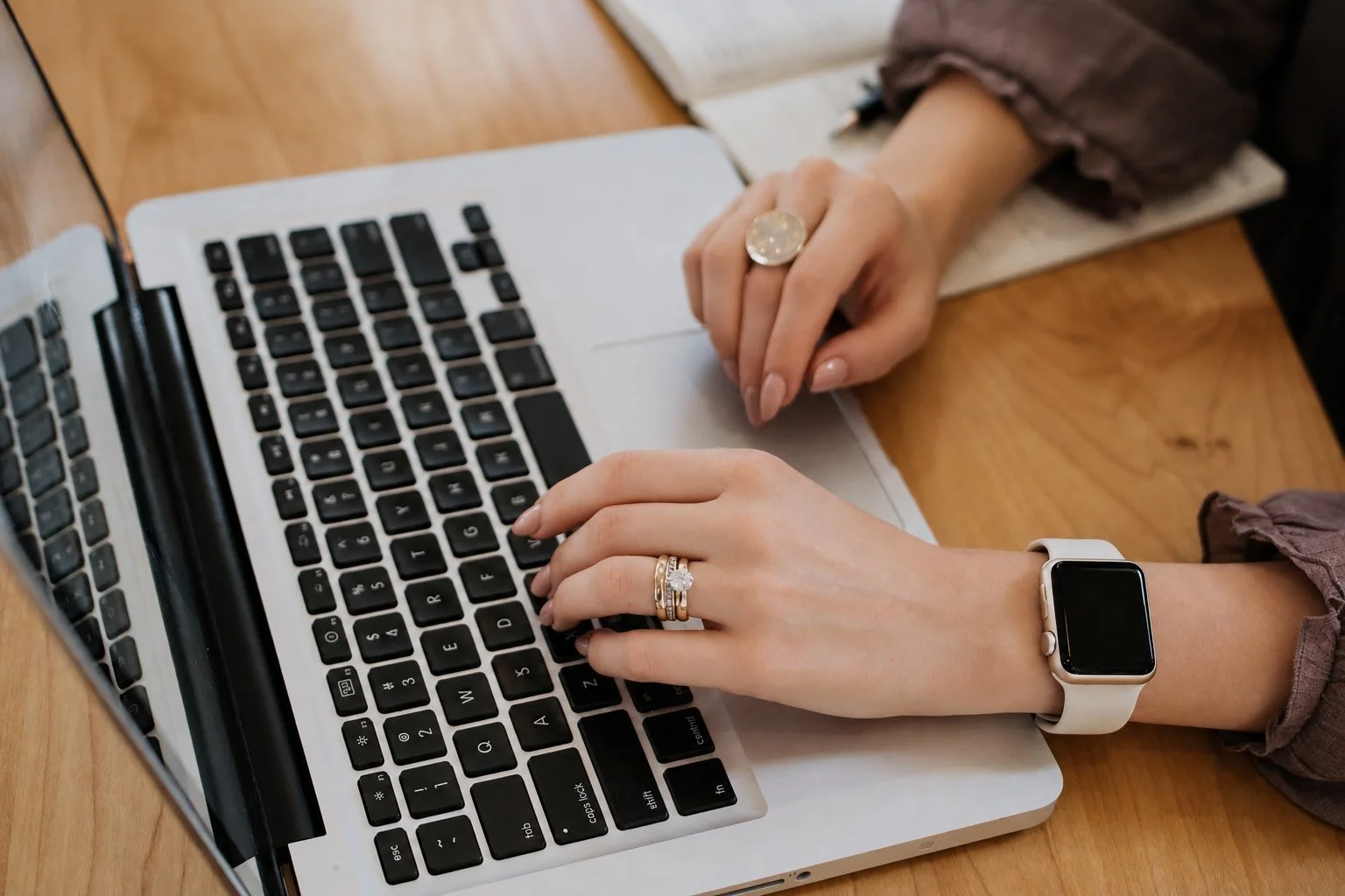 A person typing on a silver laptop with a black keyboard, wearing a smartwatch on their left wrist and multiple rings on their fingers, sitting at a wooden table.