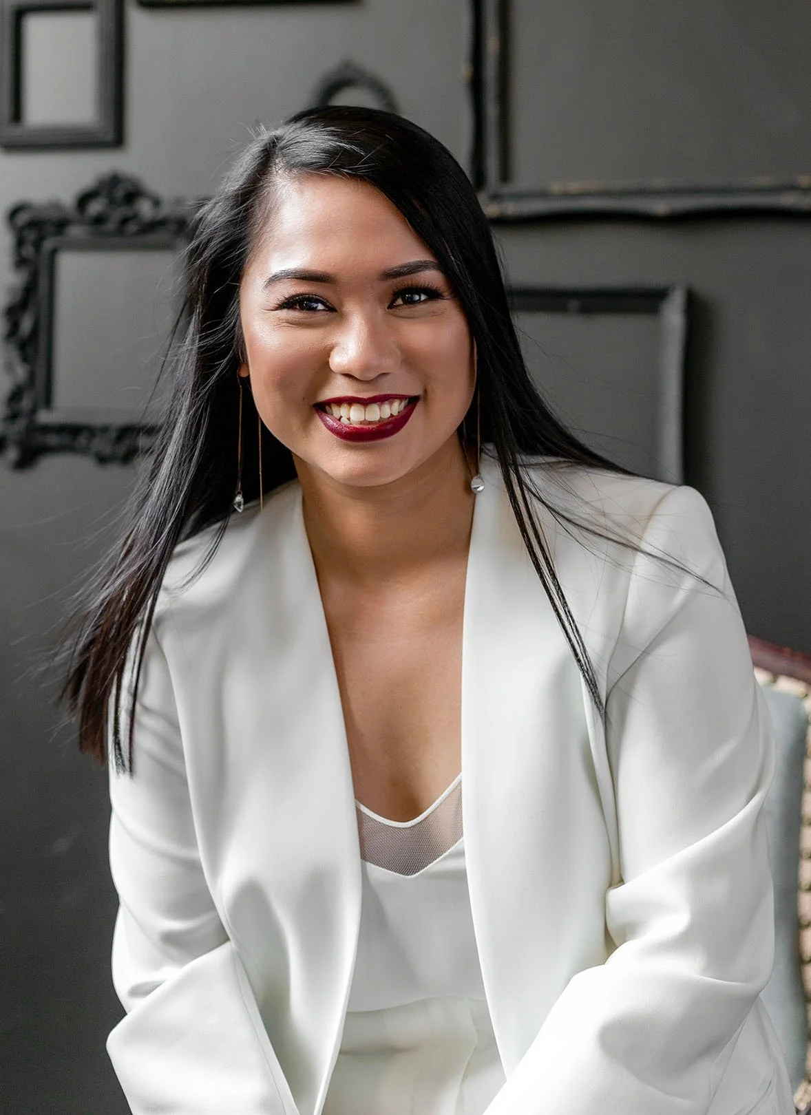 A smiling woman with long black hair, wearing a white blazer, sitting indoors with a dark background.