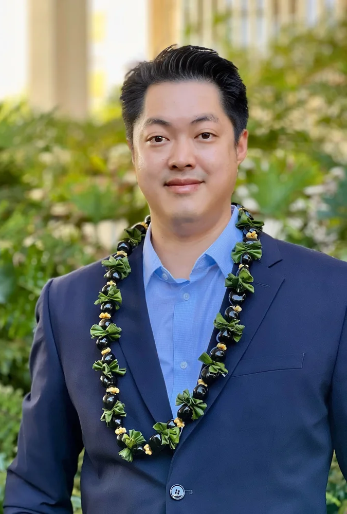 A man in a dark suit and blue dress shirt wearing a lei, standing outdoors with greenery in the background. Partially visible text in the top left corner reads 'David Kwon'.