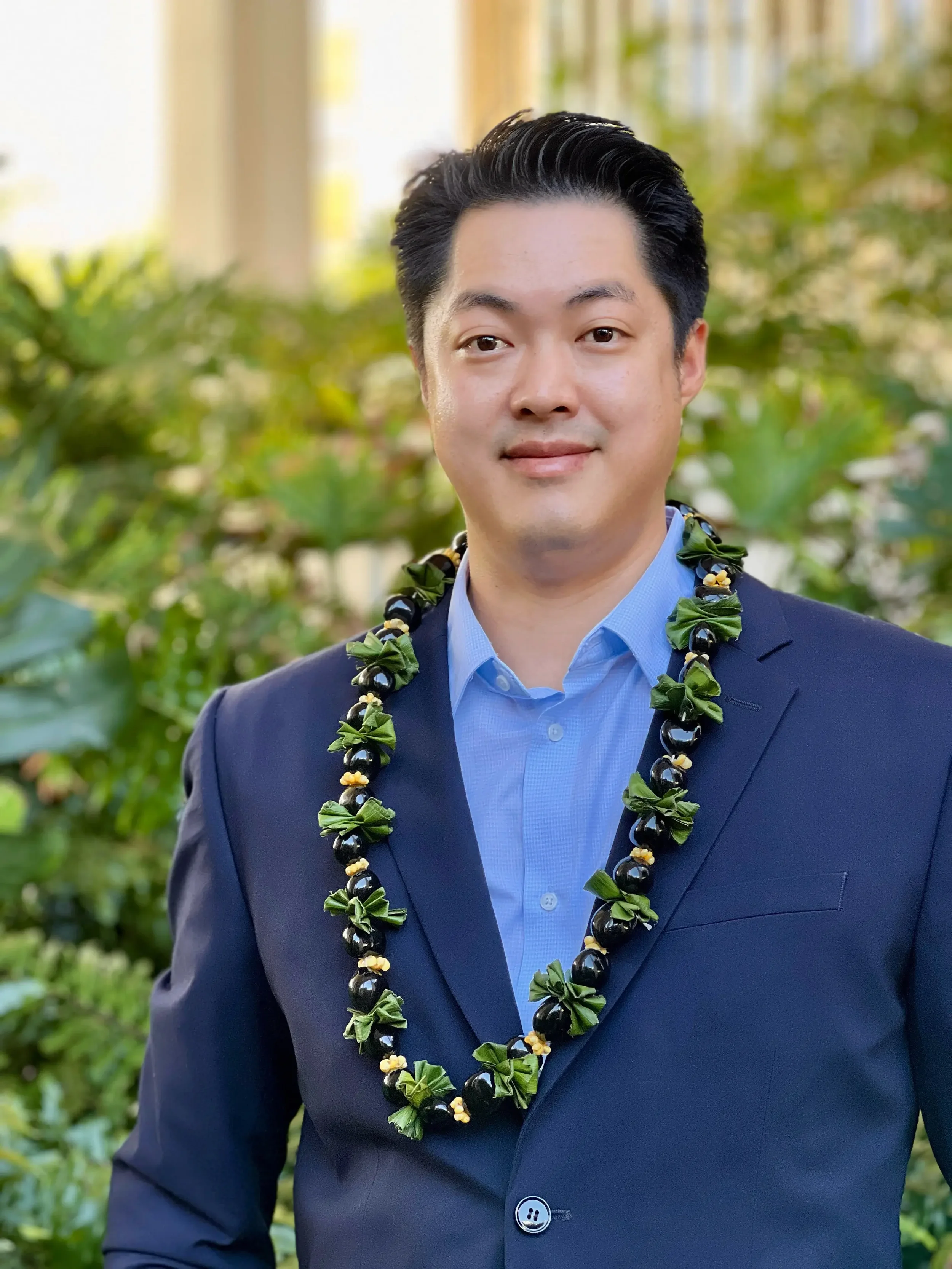 A man in a dark suit and blue dress shirt wearing a lei, standing outdoors with greenery in the background. Partially visible text in the top left corner reads 'David Kwon'.