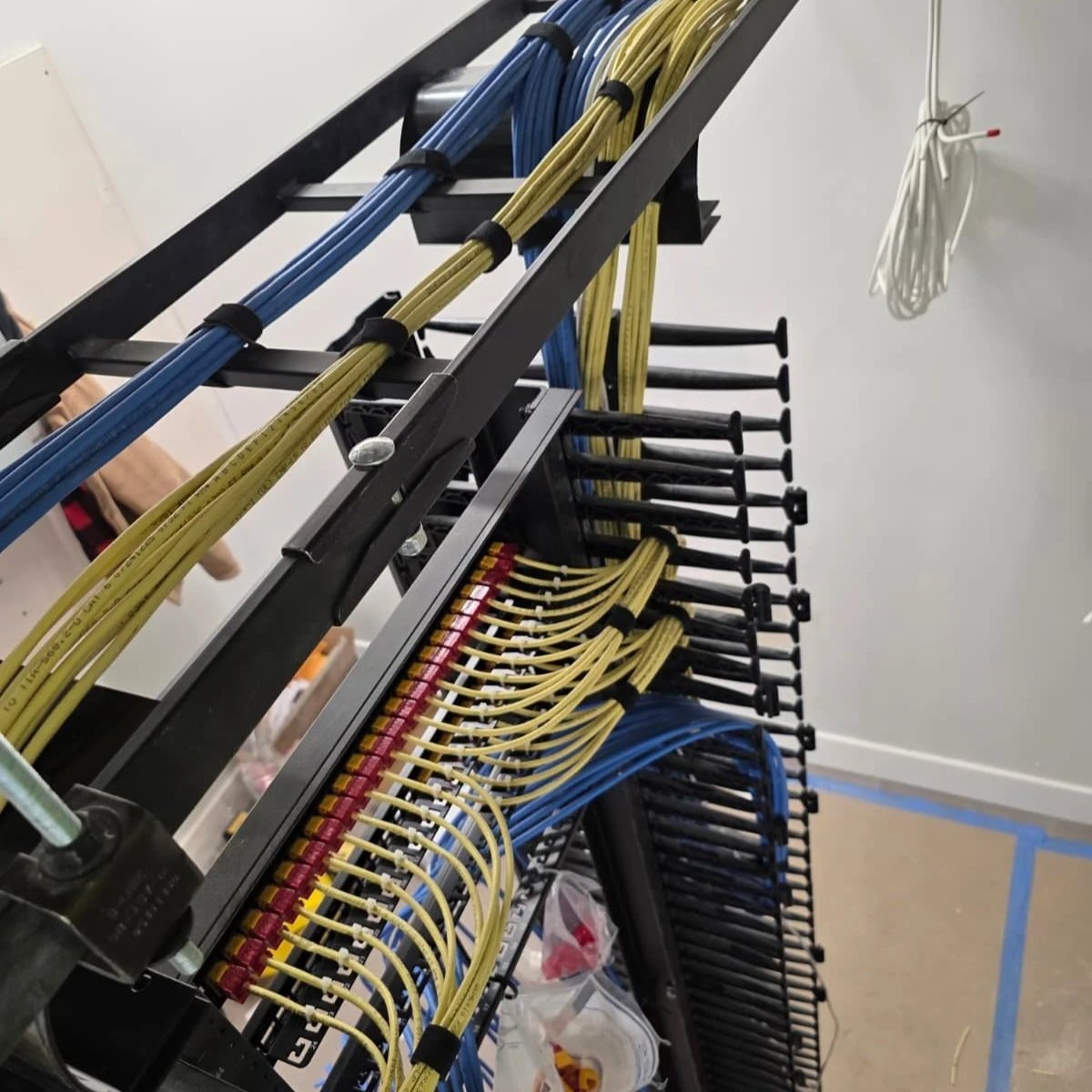 Network patch panel with yellow and blue Ethernet cables, mounted on a black metal frame in a server room.