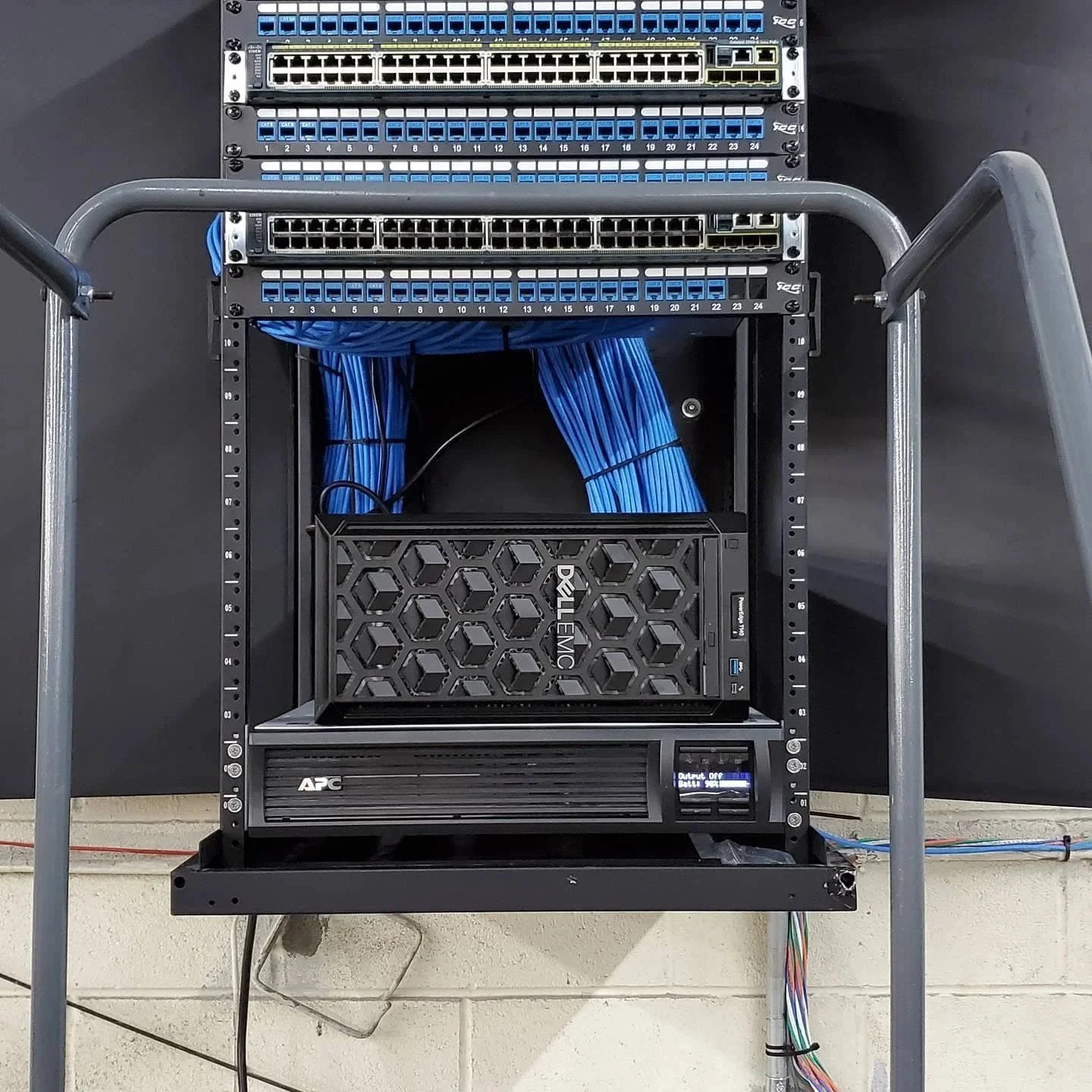 A network rack with multiple switches, a Dell EMC device, and a power distribution unit, with blue Ethernet cables organized inside, mounted on a cart against a black wall.