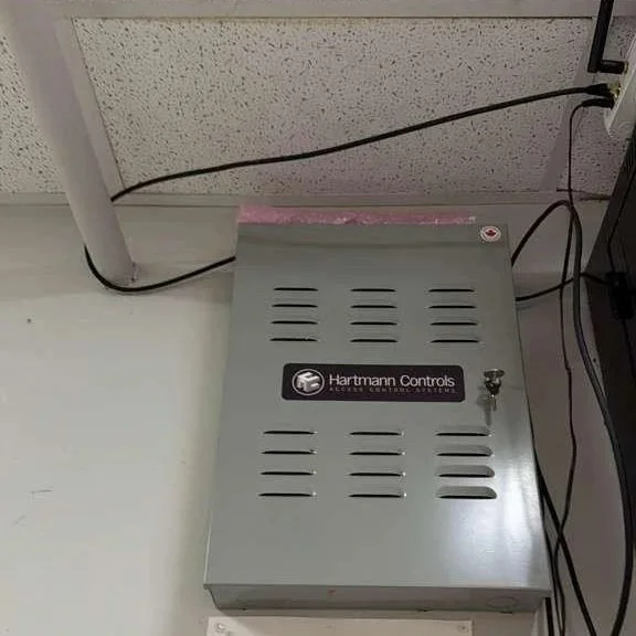 Metal electrical control box with a label saying "Hartmann Controls" and various vents, connected to black and white cables.
