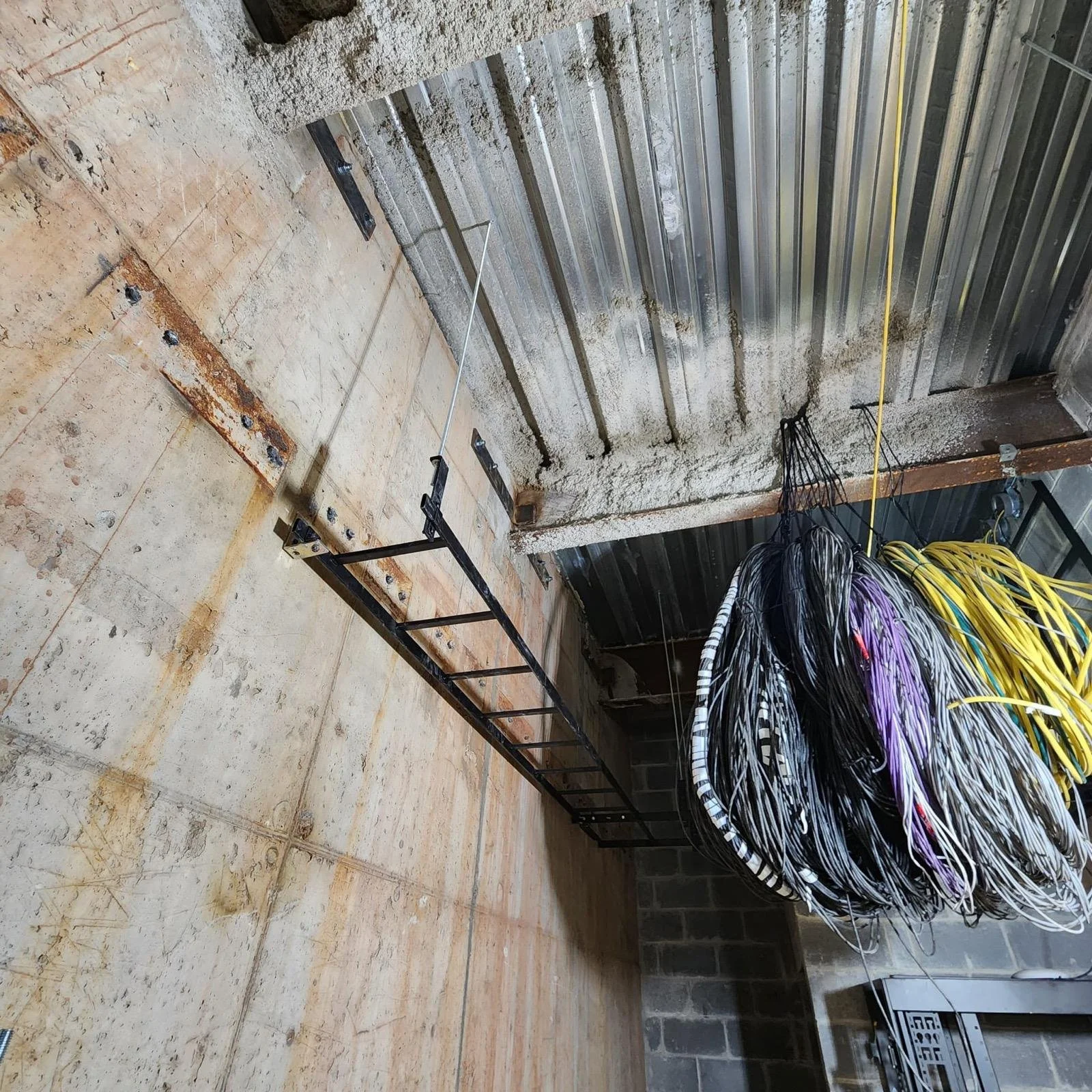 A construction site with exposed concrete floors and walls, a metal support beam, and a large bundle of black, white, purple, and yellow cables hanging down. Yellow electrical wires run along the ceiling.