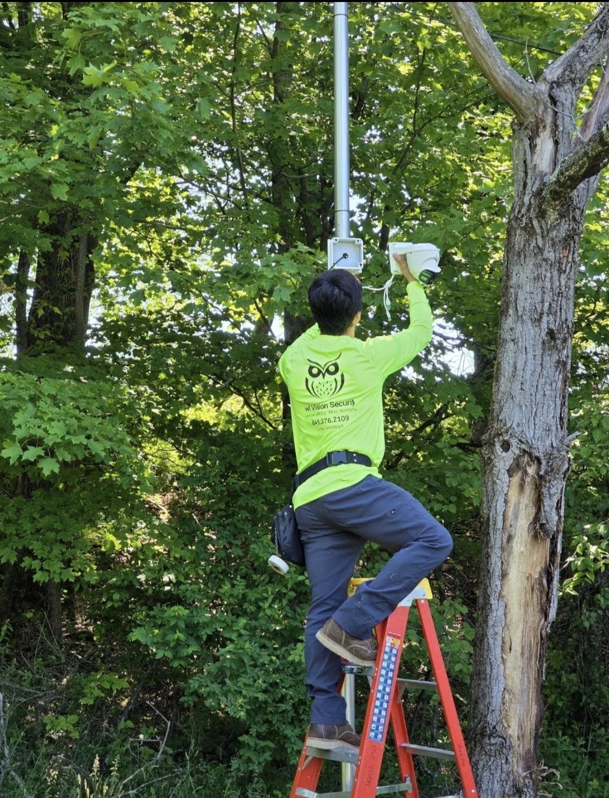 A worker wearing a yellow-green shirt with the logo 'W Vision Security' and blue pants standing on a red ladder, installing a security camera on a metal pole amidst green trees in a forested area.