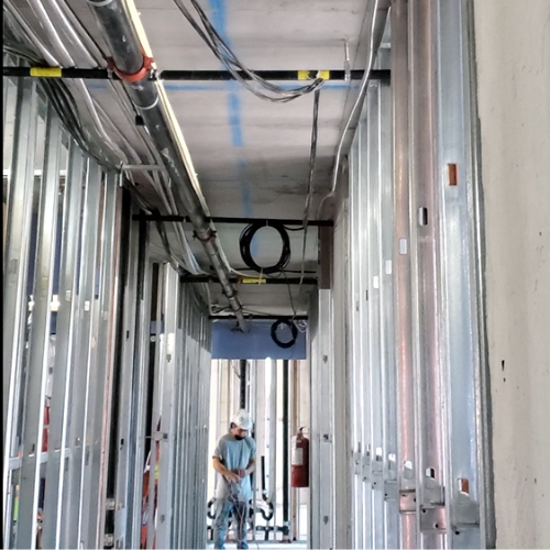 Construction worker in a partially built hallway or corridor with metal framing and exposed electrical conduits and pipes.