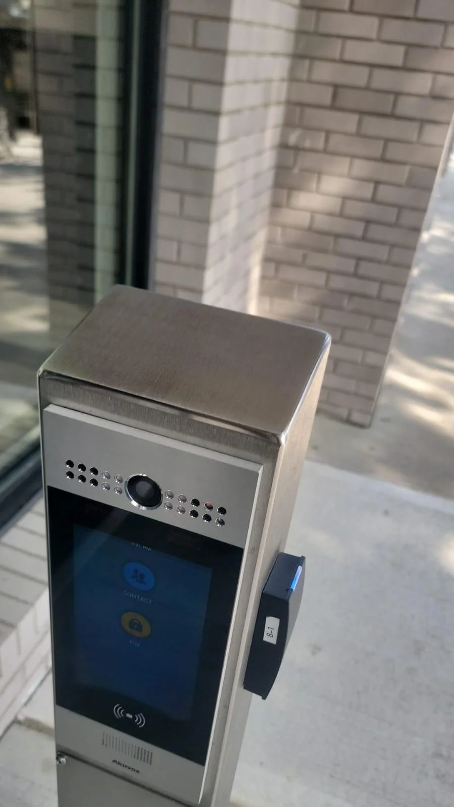 Touchless building access control panel with contactless and camera features outside a building with brick walls.