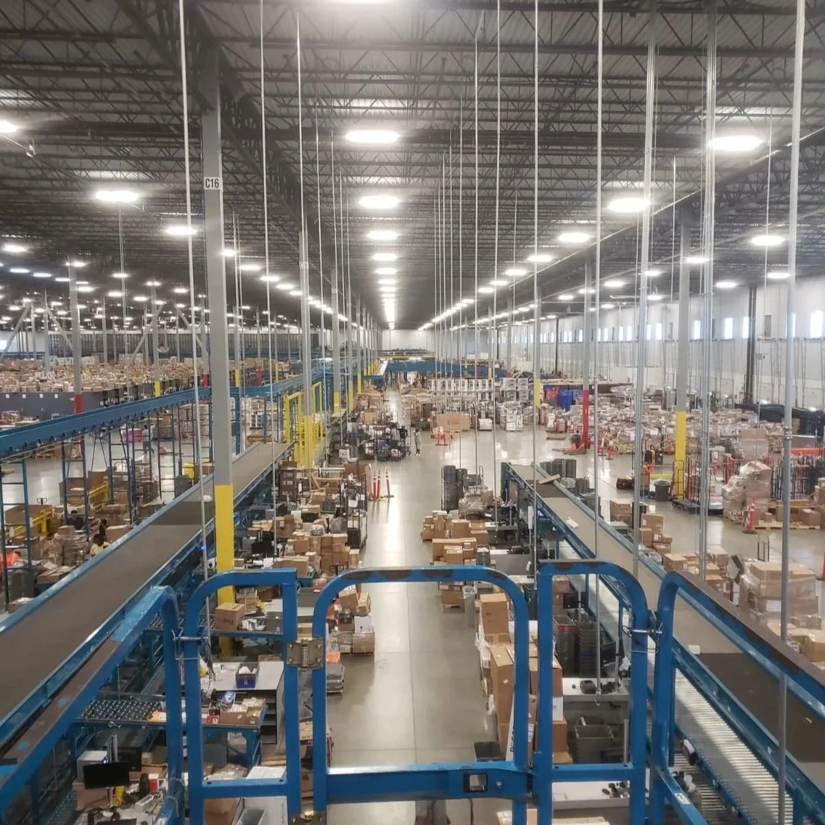 A large warehouse with rows of shelves filled with boxes and packages, illuminated by bright ceiling lights, with a blue elevated platform in the foreground.