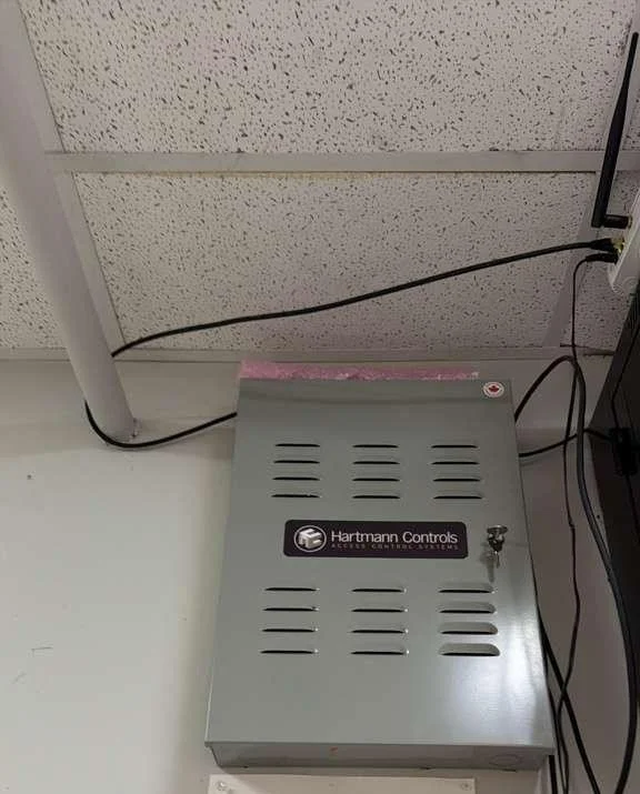 A metal control box labeled 'Hartmann Controls' on a white wall. There are cables connected to it and a ceiling with acoustic tiles above.