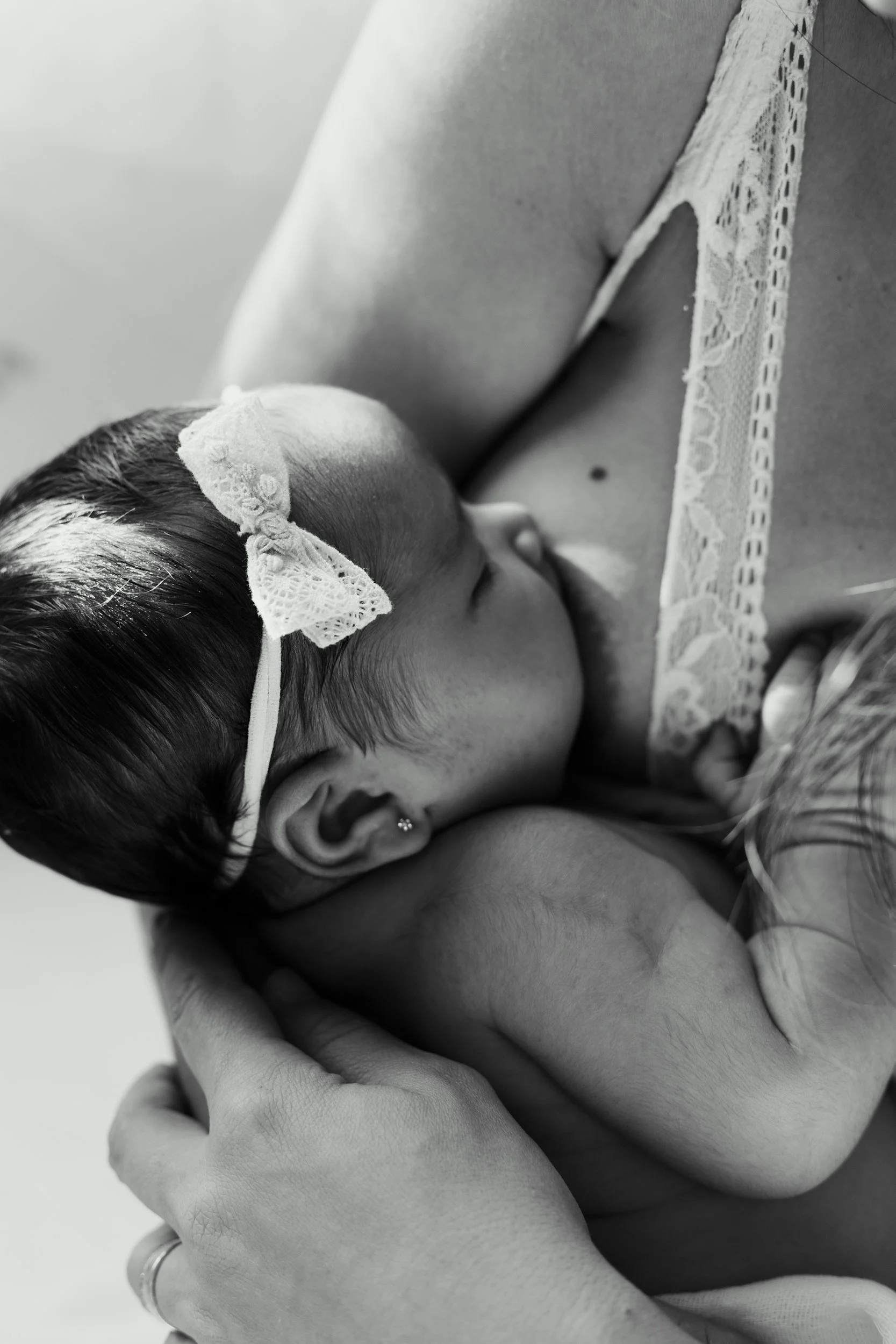 A black and white photo of a newborn baby resting on an adult's chest, wearing a lace headband with a bow.