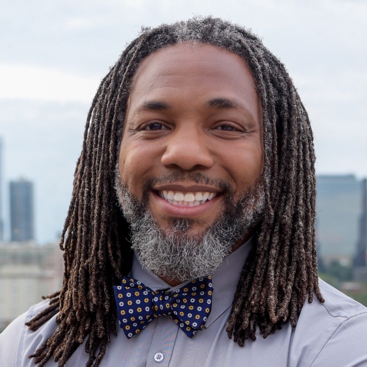 A smiling man with dreadlocks and a gray beard, wearing a light blue shirt and a blue bow tie with yellow and white patterns, outdoors with a cityscape in the background.