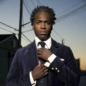 A man with dark skin and styled hair adjusting his tie outside during the day, with power lines and buildings in the background.