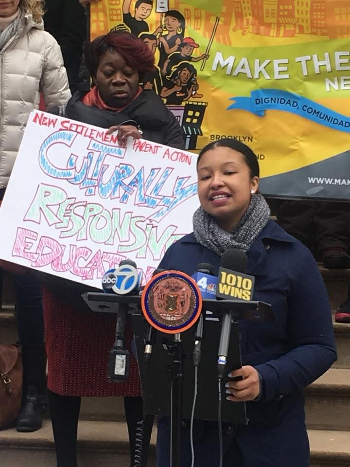 A young girl speaking at a podium during a press conference, with microphones from ABC 7, 4, and 1010 Wins. Behind her, a woman holds a sign that reads 'New Settlement Parent Action' and 'Gurrilla Responsible Education.' A colorful banner with people holding signs and a phrase related to making the news is visible in the background.