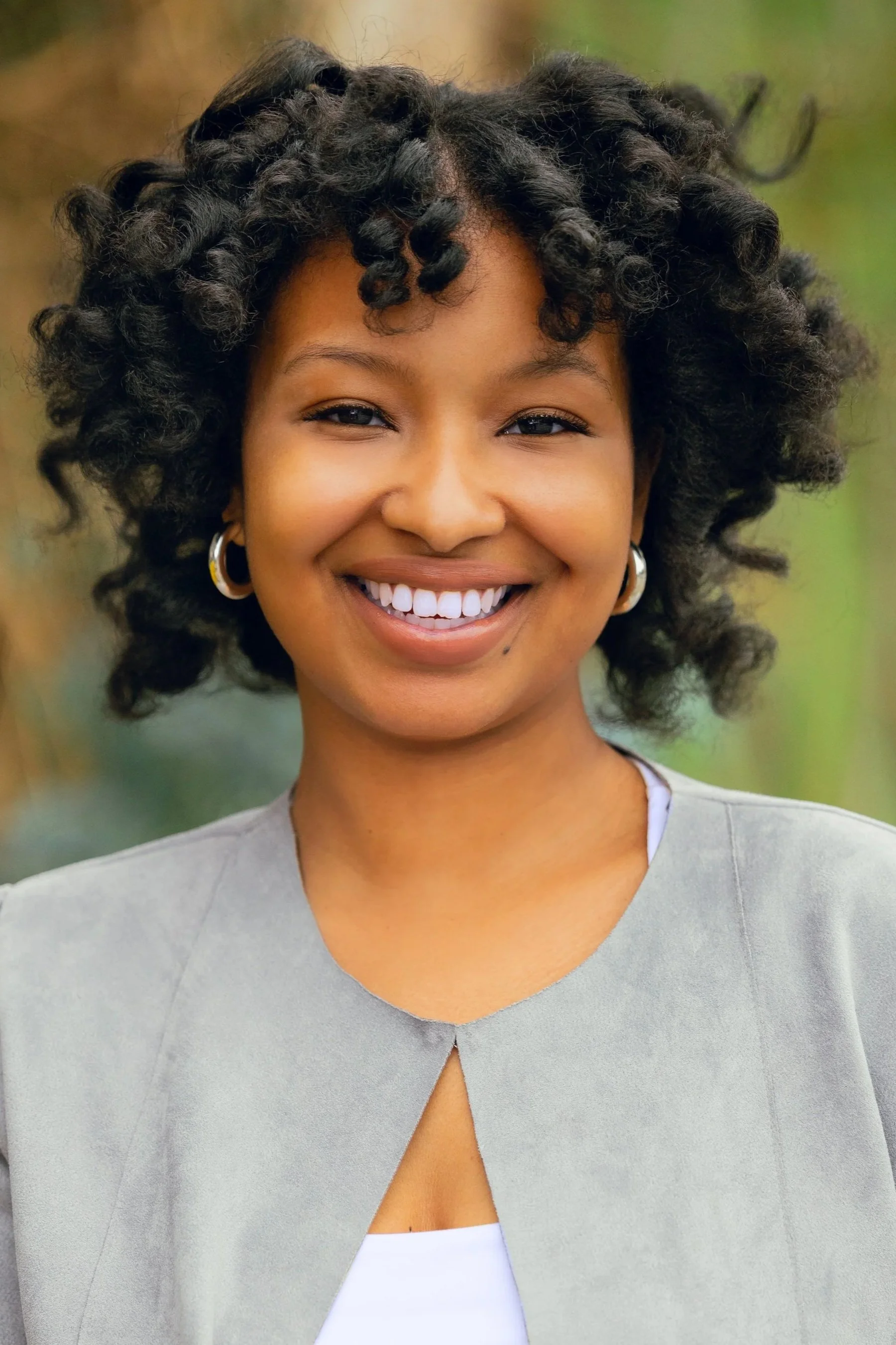 Smiling woman with curly hair wearing earrings and a light-colored jacket outdoors.