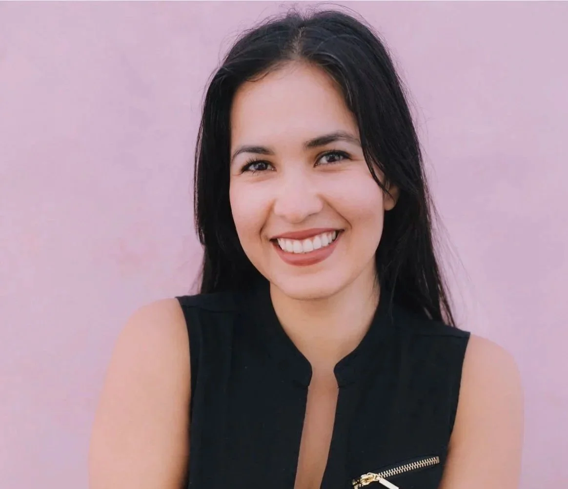 A young woman with dark hair and a big smile, wearing a sleeveless black top, poses against a light pink background.