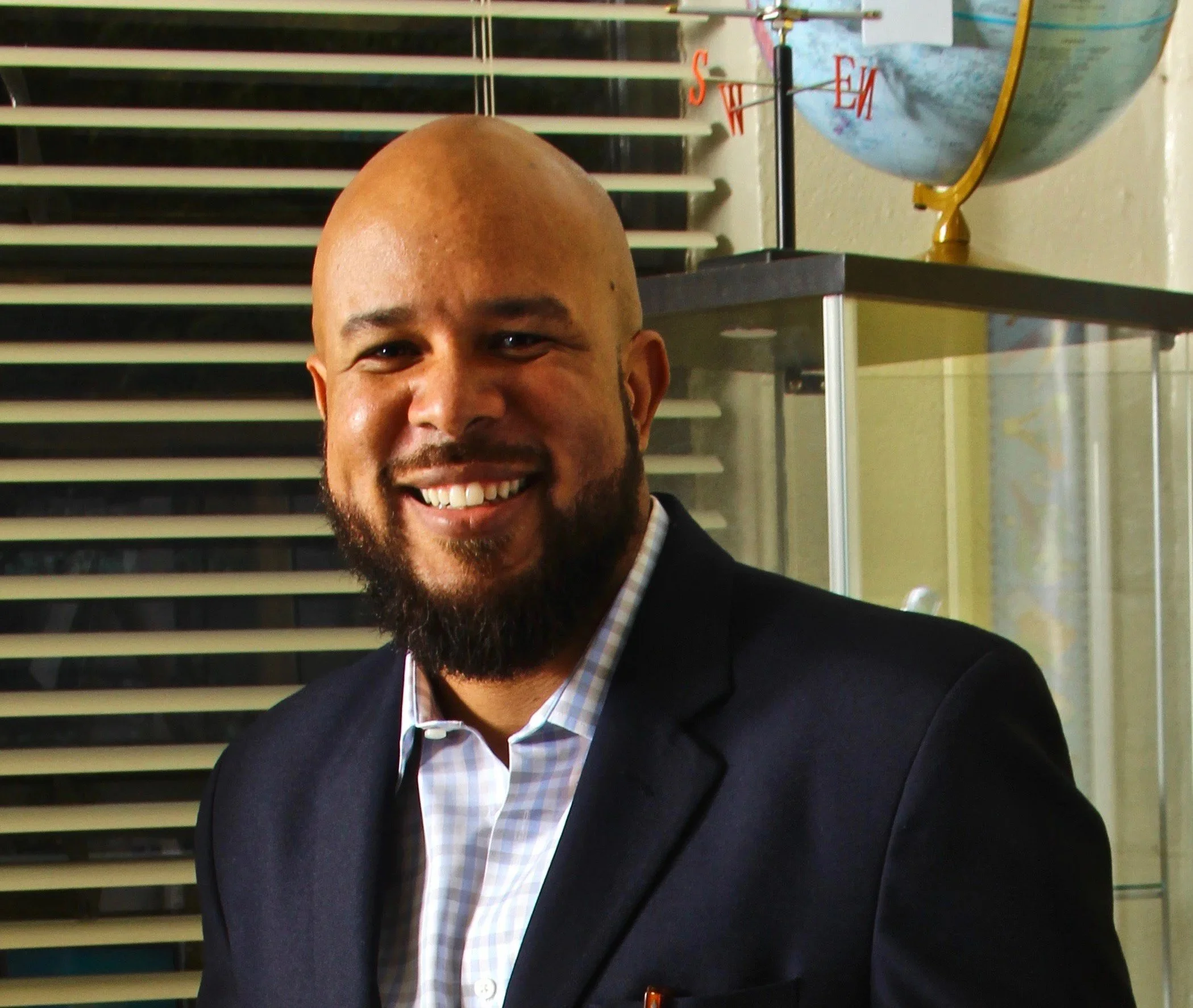 A smiling man with a beard and bald head wearing a dark suit and light checkered shirt, standing in front of a window with blinds, with a globe and a compass decoration in the background.