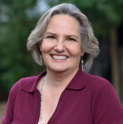 A woman with gray hair smiling outdoors, wearing a maroon blazer and a necklace.