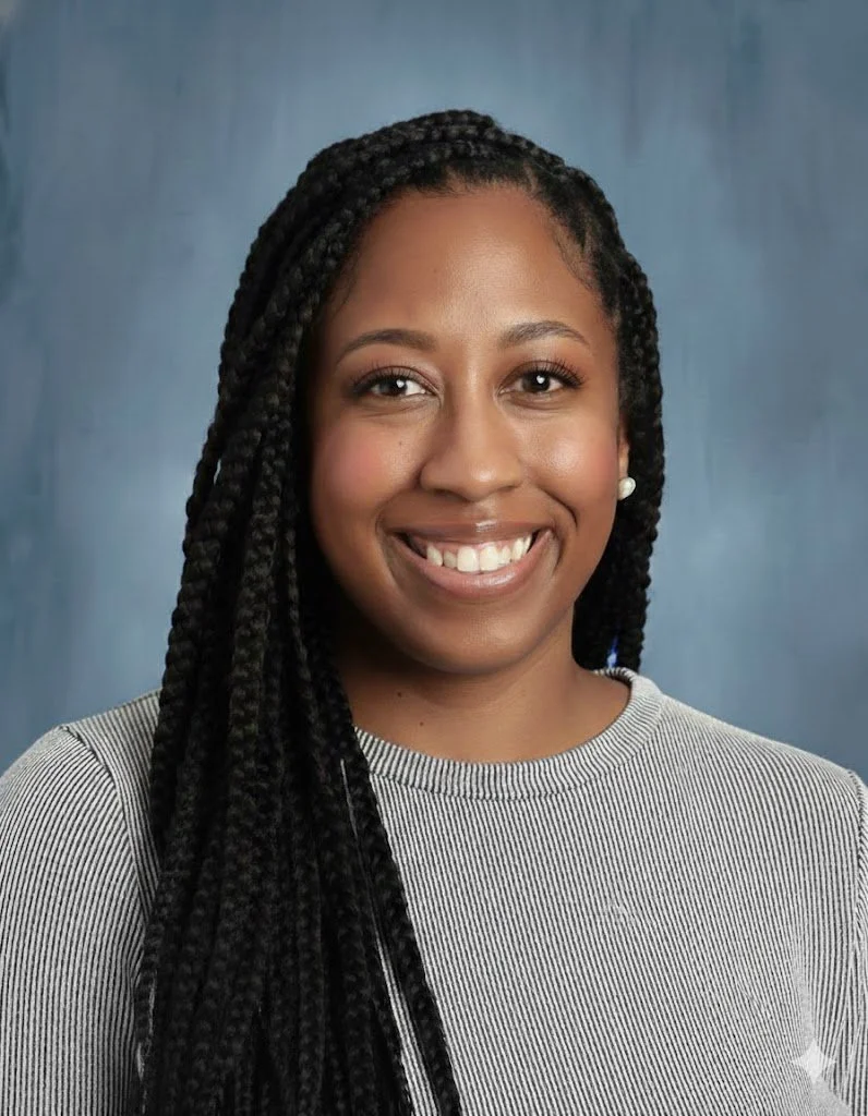 A smiling young African American woman with long braided hair, wearing a grey striped top and pearl earrings, posing against a mottled blue backdrop.