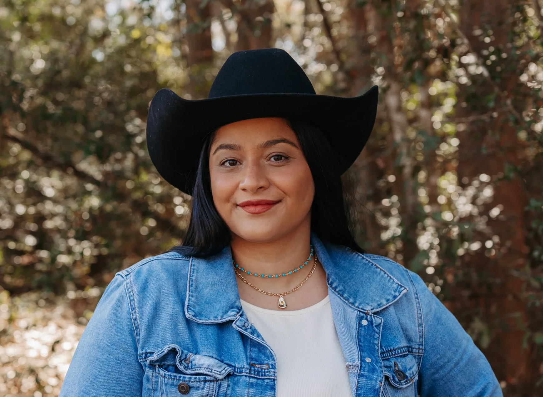 A woman in a black cowboy hat, blue denim jacket, and layered necklaces standing outdoors in front of trees.
