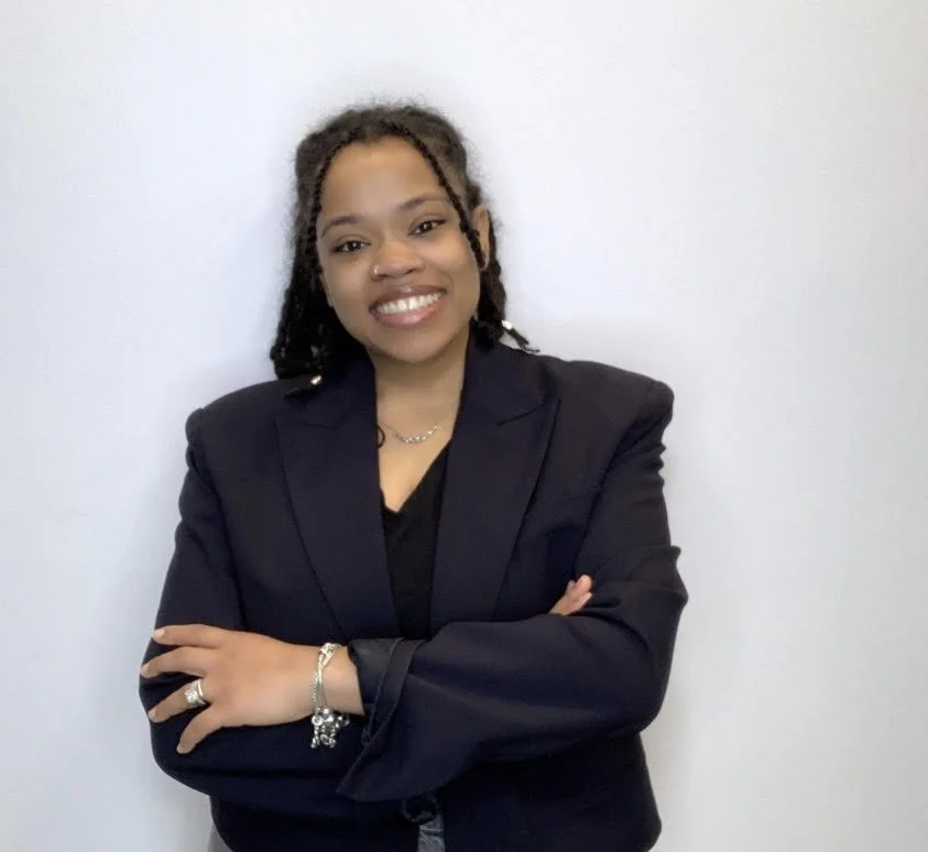 Woman with dark curly hair in a blazer, smiling with arms crossed, standing against plain light wall.
