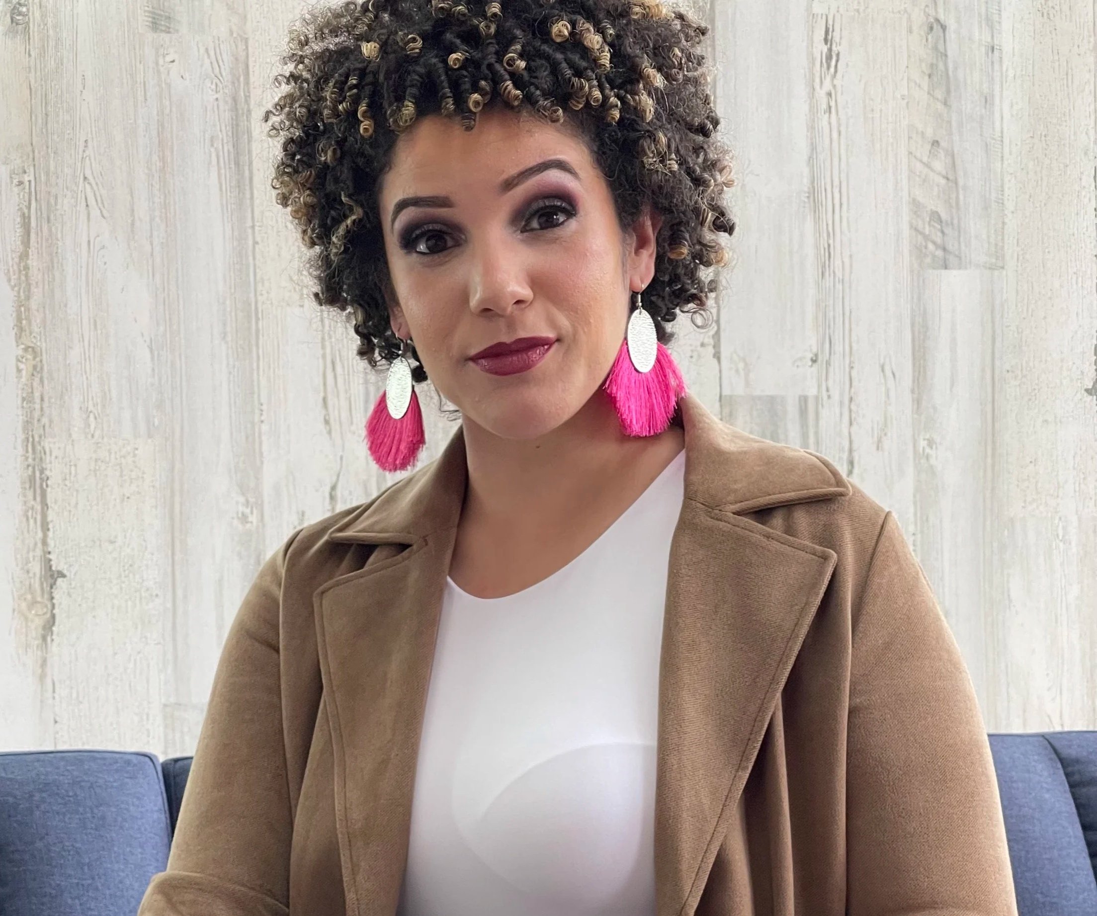 A woman with short, curly hair and dark makeup, wearing large white oval earrings with pink tassels, a white top, and a tan coat, sitting on a blue couch in front of a light-colored wooden wall.