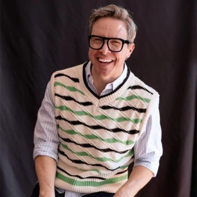 A smiling man wearing glasses, a striped sweater vest over a button-down shirt, against a dark background.