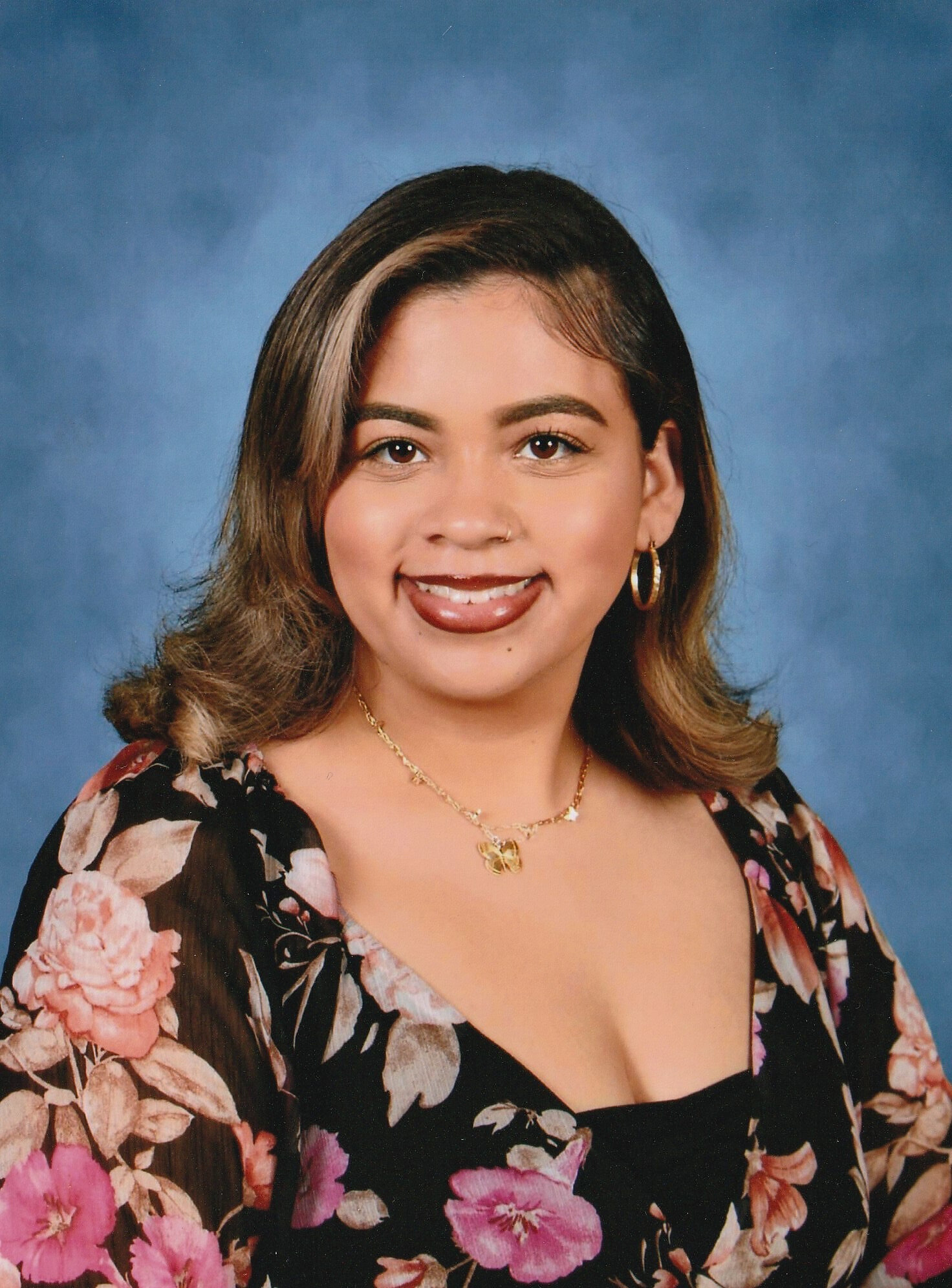 A young woman with medium-length brown hair wearing a floral top, gold jewelry, and earrings, smiling against a blue background.