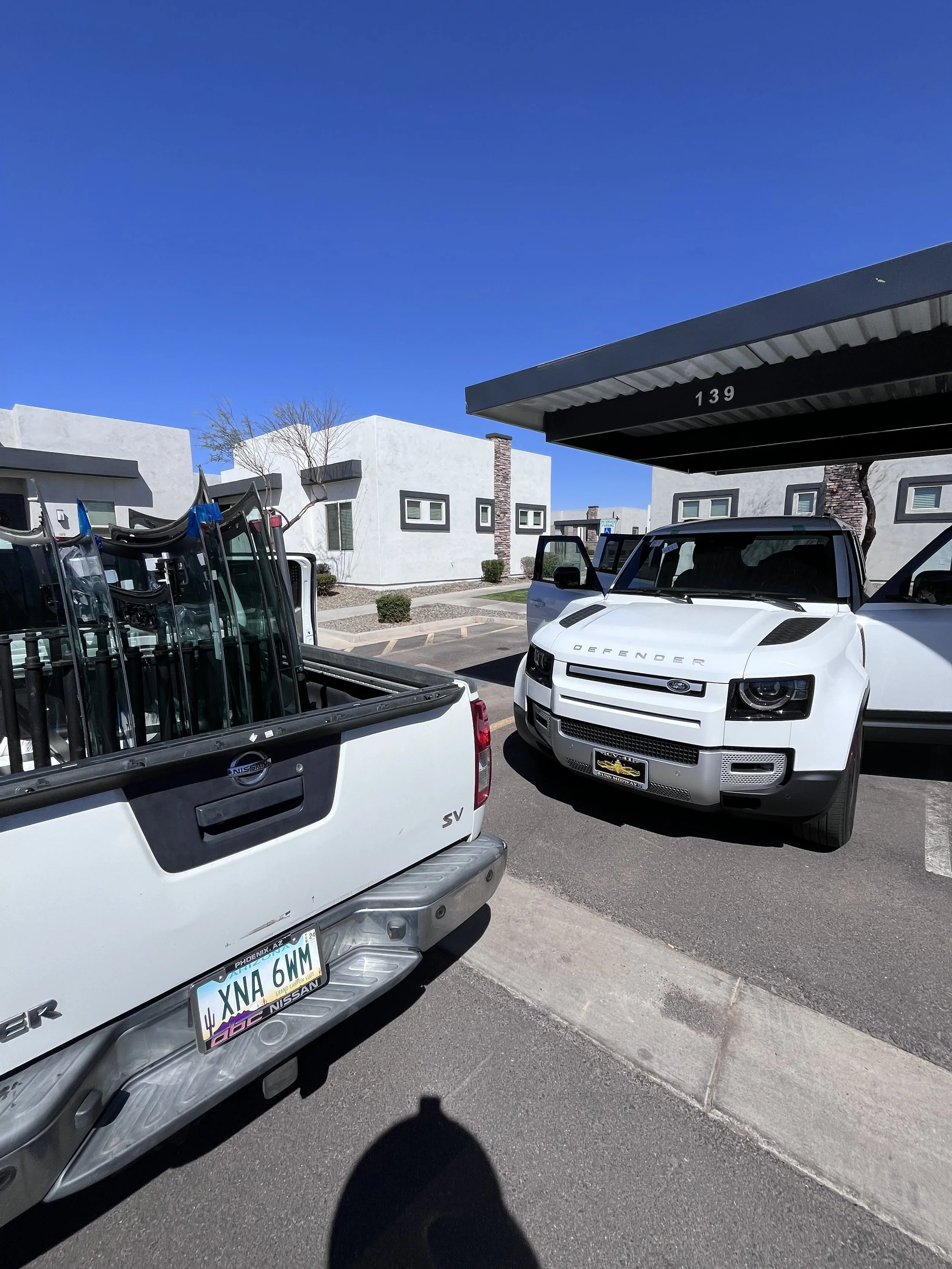 A parking lot with white pickup trucks, one labeled 'Nissan' and another labeled 'Defender', against a backdrop of modern white buildings and a clear blue sky.