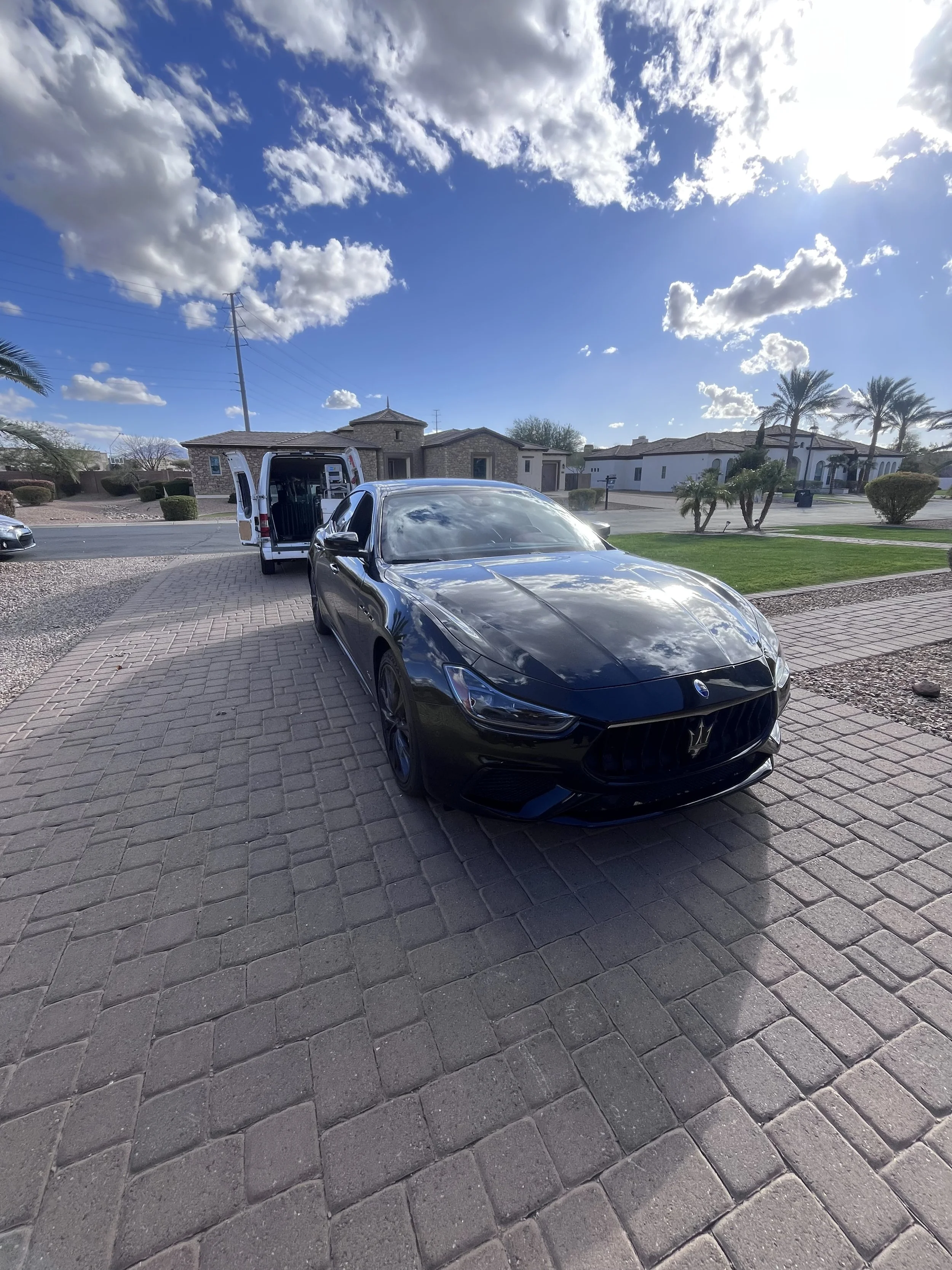Black Maserati Ghibli parked on a brick driveway with a white van behind it, in a neighborhood with palm trees and houses under a partly cloudy sky.