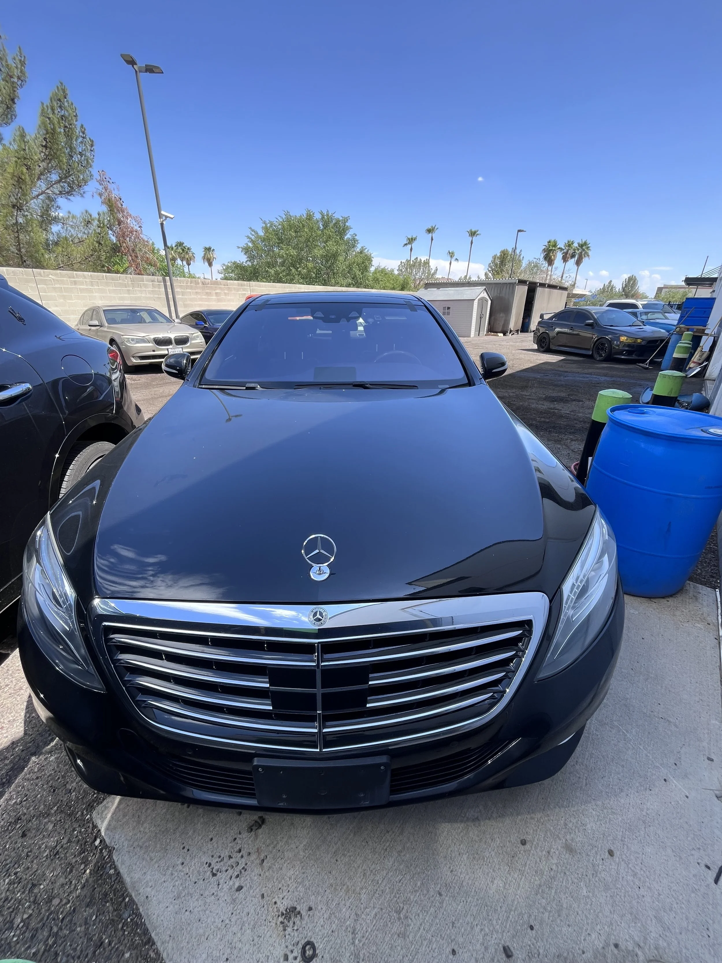 Black Mercedes-Benz car parked in a lot with other vehicles, trees, and blue barrels nearby under a clear blue sky.
